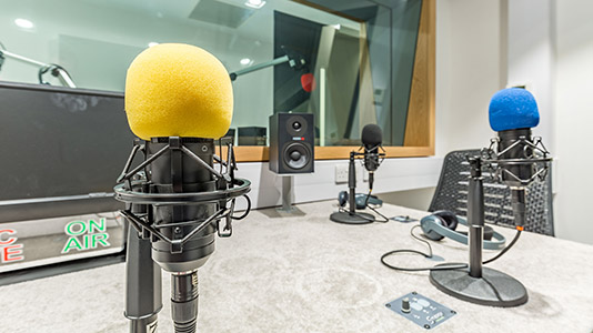Podcast recording studio set up for radio news, featuring three microphones with colored foam windscreens (yellow, black, and blue), a pair of headphones, speakers, and an "On Air" sign.
